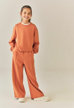 Ragazza sorridente con felpa larga color ruggine e pantaloni ampi con le mani nelle tasche, indossa scarpe da ginnastica bianche, in piedi su sfondo neutro.