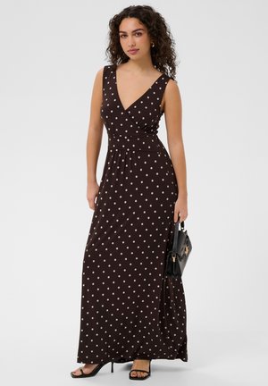 Femme aux cheveux bouclés portant une robe longue noire sans manches à pois blancs, tenant un petit sac à main noir et portant des talons noirs.
