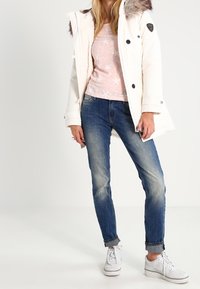 Vit parkas med avtagbar pälskrage, svarta knappar, ljusrosa skjorta med sjöstjärnemönster, blå jeans och vita sneakers.