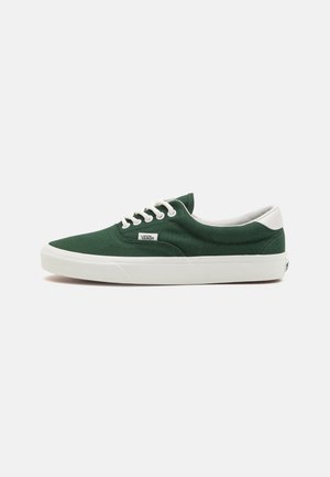 Zapatilla Vans de caña baja de color verde oscuro con cordones blancos, suela blanca y lengüeta trasera blanca sobre fondo claro liso.