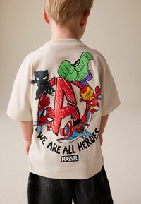 Kremasto bela bombažna majica z barvitim motivom Marvelovih superjunakov in napisom »WE ARE ALL HEROES« na hrbtni strani.
