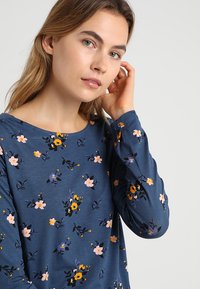 Camicia a maniche lunghe color navy con un motivo floreale in rosa, giallo e nero. Tessuto morbido, scollatura tonda e maniche aderenti.
