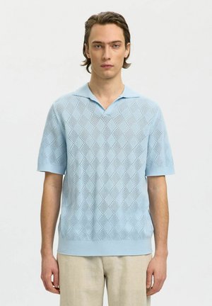 Bărbat tânăr purtând un tricou polo tricotat bleu cu mâneci scurte și un model în formă de diamant și pantaloni bej, stând în fața unui fundal simplu.