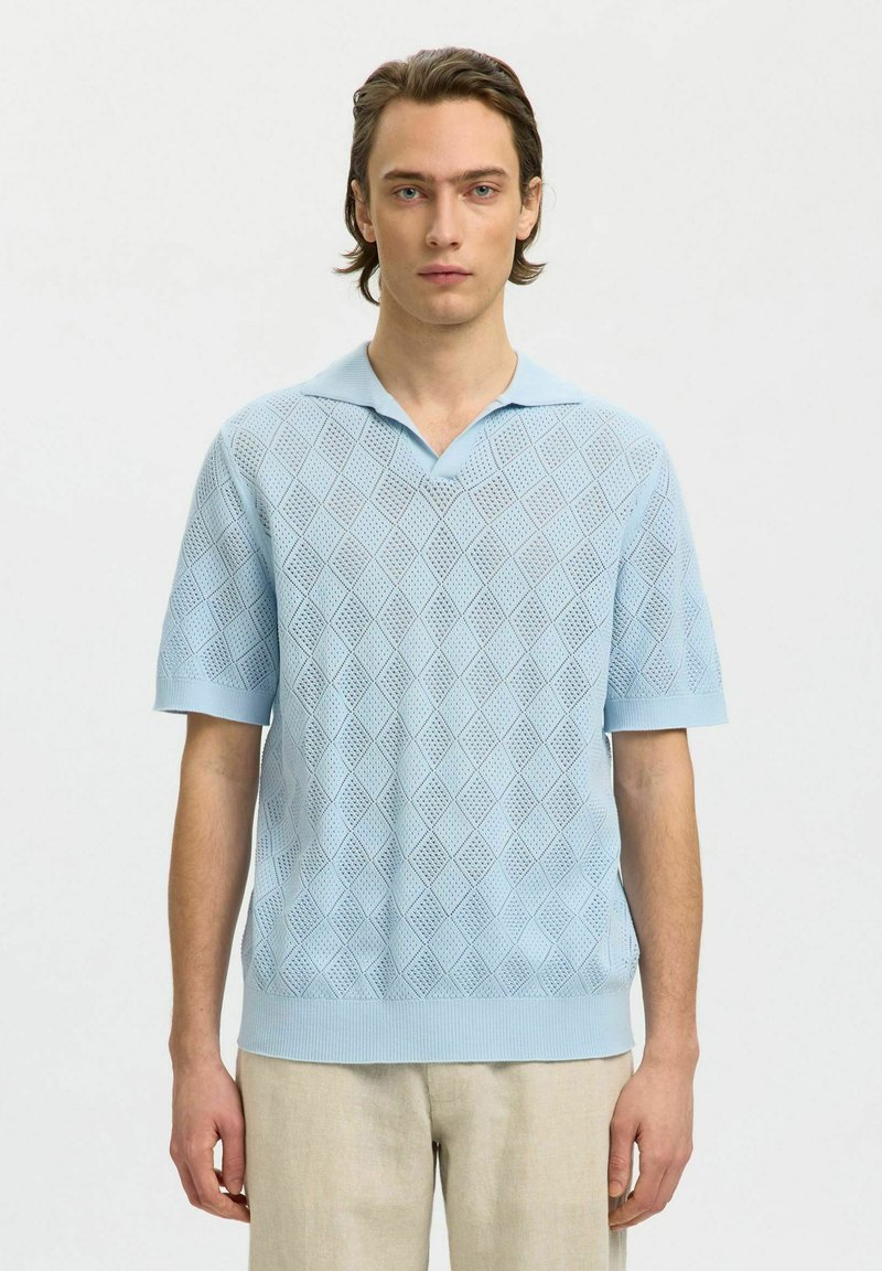 Jeune homme portant un polo en maille bleu clair à manches courtes avec un motif losange et un pantalon beige, debout devant un fond uni.