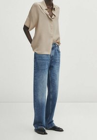 Beige overhemd met knoopsluiting, losse pasvorm en korte mouwen, gecombineerd met lichtblauwe wijde spijkerbroek. Zwarte platte sandalen maken de look compleet.