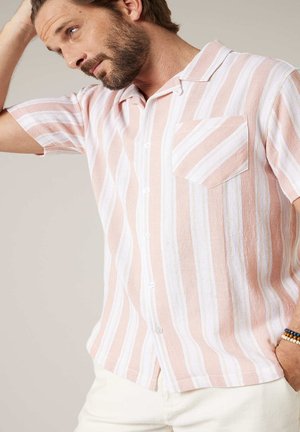 Man met baard, gekleed in een kort mouws overhemd met roze en witte verticale strepen en een borstzak, met zijn hand op zijn hoofd, tegen een effen achtergrond.