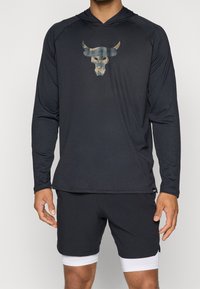 Langärmliger schwarzer Hoodie mit einem Camouflage-Bullenlogo auf der Brust. Hergestellt aus glattem, dehnbarem Material. Kombiniert mit schwarzen Sportshorts.