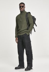 Suéter de punto acanalado verde con cremallera en el cuello, pantalones cargo negros con bolsillos laterales, botas negras y una mochila negra. Incluye gafas de sol oscuras.