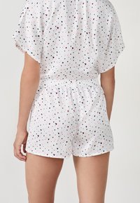 Mujer vestida con pantalones cortos y top blancos con pequeños patrones de corazones en rosa, azul marino y burdeos, vista desde la espalda contra un fondo liso.
