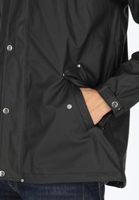 Czarna wodoodporna kurtka wykonana z gładkiej, teksturowanej tkaniny. Posiada srebrne zatrzaski i boczne kieszenie z przeszytymi krawędziami.