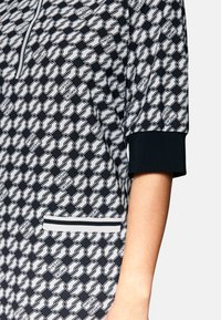 Kurzärmliges Oberteil mit schwarz-weißen Mustern, Reißverschlusskragen und gerippten Bündchen, ausgestattet mit einer Fronttasche mit gestreiftem Detail.