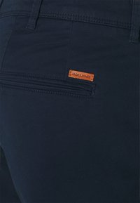 Tamno plave hlače imaju teksturiranu tkaninu i stražnji džep. Mali narančasti kožni oznaka s natpisom "JACK & JONES" ušivena je iznad džepa.