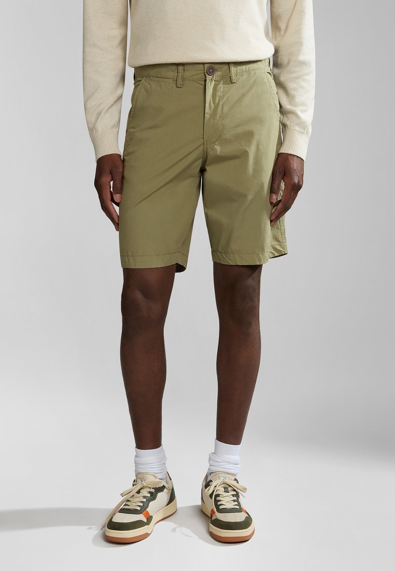 Olijfgroene shorts van lichtgewicht stof, met een knoopsluiting en zijzakken. Gepaard met crèmekleurige sneakers en witte sokken.