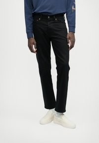 Črne kavbojke s ravnim krojem, narejene iz denima, z oranžnim šivanjem in klasičnim petim žepom, v kombinaciji z belimi natikači.