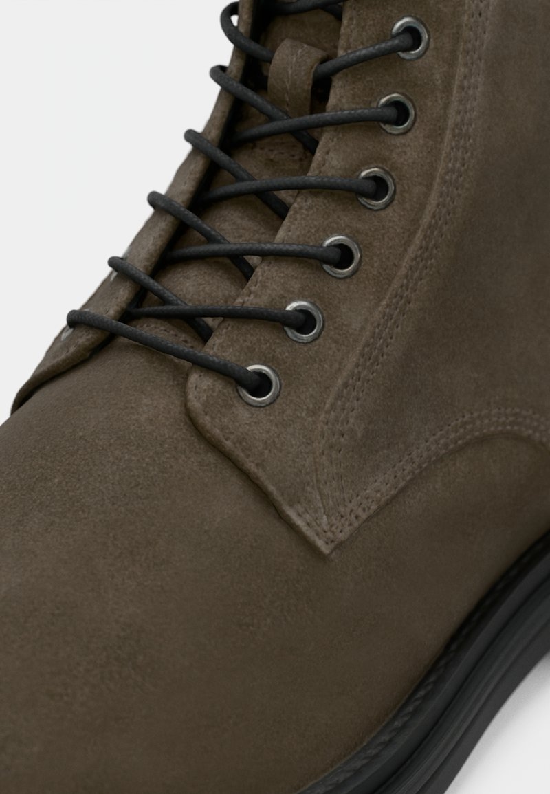 Botín de ante marrón con cordones negros, ojales de metal y una superficie texturizada. Presenta una punta redondeada y una suela de goma negra.