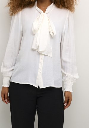 Kvinde iført en hvid bluse med lange ærmer, bindebånd foran i halsen og smockede manchetter, kombineret med sorte bukser, stående mod en ensfarvet baggrund.