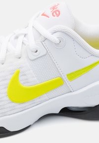 Zapatilla deportiva blanca con parte superior de malla, el logotipo de Nike en amarillo y detalles, que presenta cordones y un detalle del logo en rosa claro en la lengüeta.