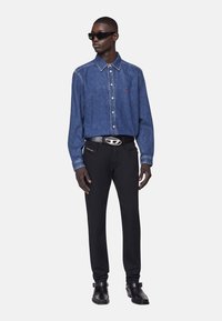 Man med mörka solglasögon, blå denimskjorta, svart bälte med silverspänne, svarta byxor och svarta läderskor mot vit bakgrund.