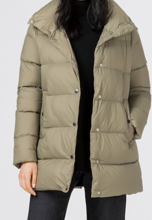 Personne portant une doudoune matelassée marron clair avec des boutons-pression et des poches, associée à un haut noir et un pantalon foncé.
