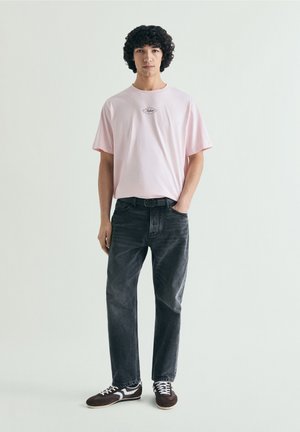 Joven con pelo rizado que lleva una camiseta rosa claro, jeans negros y zapatillas oscuras, de pie con una mano en el bolsillo.