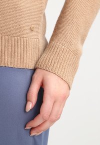 Maglione beige lavorato a maglia con polsini a coste, vista ravvicinata di una mano appoggiata su un morbido tessuto blu, che mette in mostra la texture e il dettaglio dei bottoni.