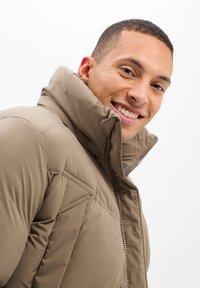 Chaqueta acolchada beige con cuello alto, cierre frontal con cremallera y detalles de costura diagonal, hecha de un tejido suave y brillante.