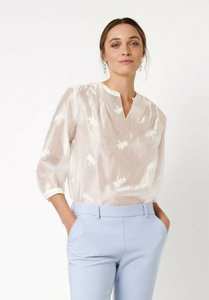 Woman wearing a sheer white blouse with floral embroidery tucked into light blue high-waisted trousers, hands in pockets, neutral background.