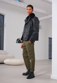 Veste noire en cuir croisé avec un col en fourrure, pantalon cargo olive et bottes noires à lacets ; texture lisse et coupe structurée.