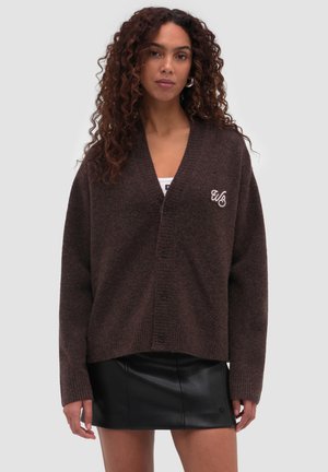 Vrouw met krullend haar draagt een donkerbruine gebreide cardigan met knopen en een zwarte leren rok, staand tegen een effen lichte achtergrond.