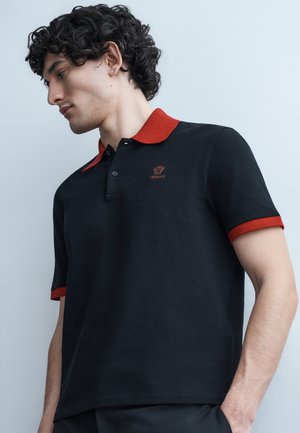 Νεαρός άνδρας με σγουρά μαλλιά που φοράει μαύρο πόλο με κόκκινο γιακά και μανσέτες, κοιτάζοντας προς τα κάτω μπροστά σε απλό φωτεινό φόντο.