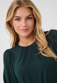 Blouse vert foncé avec encolure froncée et manches ballon. Texture lisse, tissu léger et subtil éclat qui rehaussent le design.