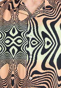 Chemise à manches courtes présentant un motif géométrique audacieux en noir et crème sur un fond pêche ; comprend un col pointu et des boutons.