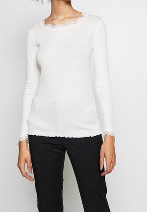 Mujer con una blusa blanca de manga larga con ribete de encaje en el escote y los puños, combinada con pantalones negros, sobre un fondo liso.