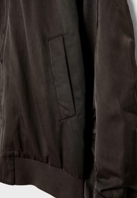 Gros plan sur la manche d'une veste en tissu noir et la poche latérale avec rabat rectangulaire cousu et ceinture élastique côtelée.