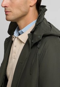 Chaqueta verde oscuro con un alto cuello, que incluye botones y cordones. Llevada sobre un polo de punto beige y una camisa azul clara.