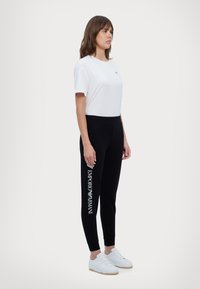 Vitt bomullströja, svarta tight sweatpants med "EMPORIO ARMANI" tryckt i vitt, bärs med vita sneakers. Släta texturer, avslappnad stil.