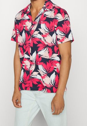 Uomo che indossa una camicia a maniche corte con bottoni, con motivo floreale rosso, bianco e rosa su sfondo scuro, abbinata a jeans azzurri.