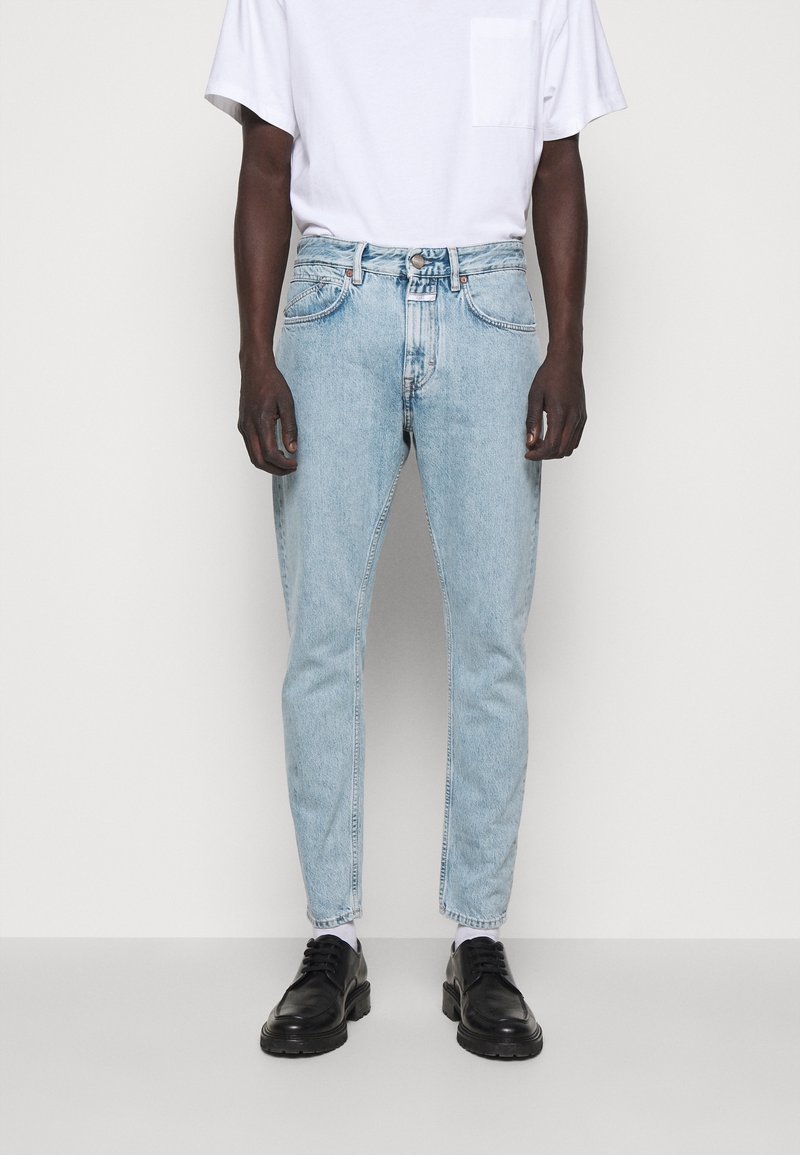 Jeans de mezclilla en un azul claro con un diseño ajustado, cinco bolsillos y costuras visibles. Combinados con zapatos negros y una camiseta blanca.