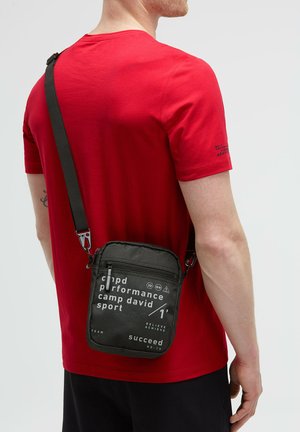 Schwarze Umhängetasche aus strukturiertem Material. Mit weißem Drucktext und einem Reißverschluss. Verstellbarer schwarzer Tragegurt und metallene Hardware-Details.