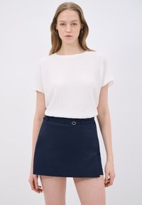 Top bianco testurizzato con maniche corte abbinato a una gonna mini blu navy con cintura e dettagli in oro.