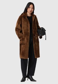 Cappotto in finta pelliccia marrone con colletto, chiusura a bottoni, due tasche laterali, indossato sopra una maglia nera e jeans scuri a vita alta. Borsa nera.