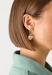 Orecchino a cerchio in oro con un pendente in vetro trasparente, con accenti turchesi e dettagli intricati. Lo sfondo mostra un colletto verde menta.