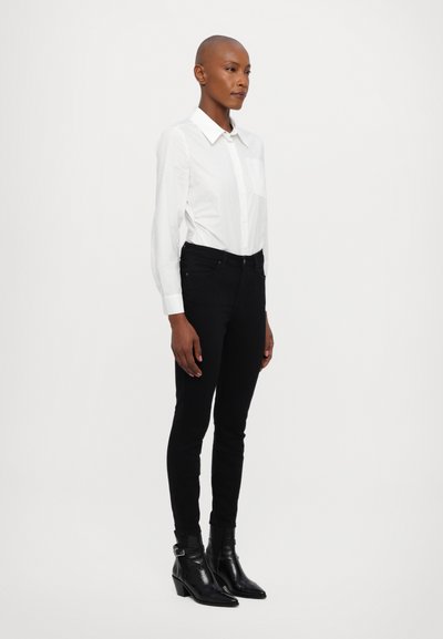 Camisa blanca de manga larga con botones y bolsillo en el pecho, combinada con jeans negros ajustados y botas negras de tobillo con un pequeño tacón. Diseño simple y elegante.