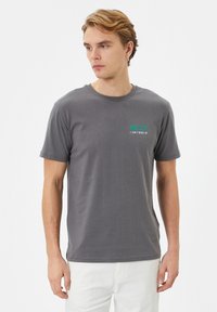 Grå bomulds t-shirt med korte ærmer, der har teksten "HJÆLP JEG KAN IKKE Vågne OP" i turkis på venstre brystområde. Glat tekstur.