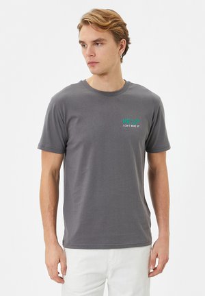 Grå bomulds t-shirt med korte ærmer, der har teksten "HJÆLP JEG KAN IKKE Vågne OP" i turkis på venstre brystområde. Glat tekstur.