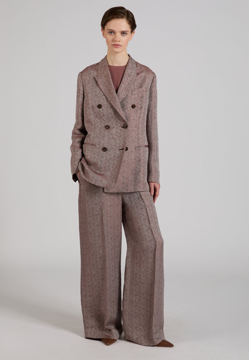 Modella che indossa una giacca doppiopetto color malva con pantaloni larghi abbinati e scarpe marroni appuntite, in piedi davanti a uno sfondo grigio uniforme.