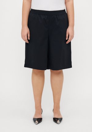 Femme portant un short noir mi-long avec une taille élastique, associé à des chaussures noires à bout pointu avec détail métallisé.