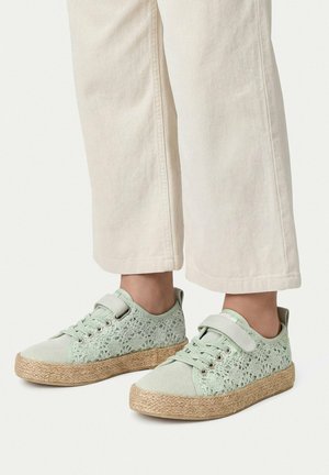 Stopy noszące jasnozielone sneakersy z koronki szydełkowej z podeszwą z juty i paskiem na rzep, zestawione z szerokimi, kremowymi spodniami.