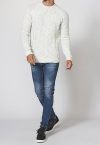 Krämfärgad stickad tröja med texturmönster i diamant, rund halsringning, ribbade ärmslut, kombinerad med blå slim-fit jeans och svarta sneakers.