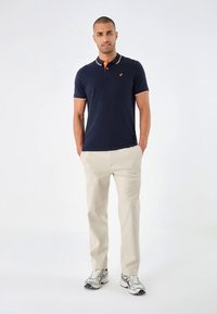 Navy polo krekls ar oranžiem apkakles akcentiem, šaura piegriezuma, kombinēts ar gaiši bēšām biksēm un pelēkām sporta kurpēm. Vienkāršs, tīrs dizains.